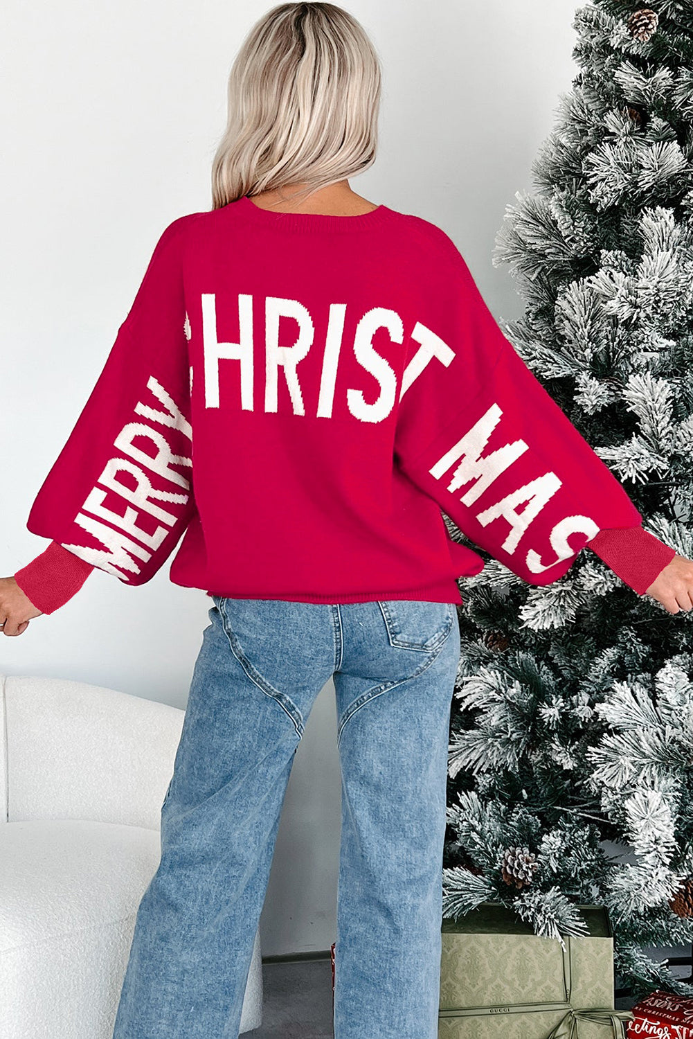 Person wearing a red 'Merry Christmas' sweater standing next to a decorated Christmas tree.