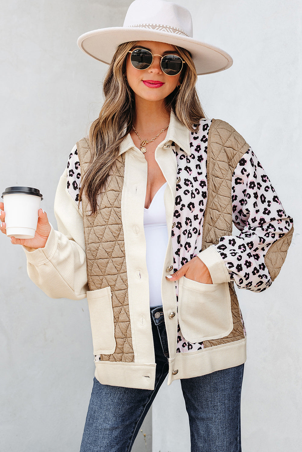 Woman wearing a leopard print and beige quilted jacket with a white hat and sunglasses, holding a coffee cup.