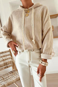 Person wearing a beige hoodie and white pants with jewelry, sitting on a wooden chair.
